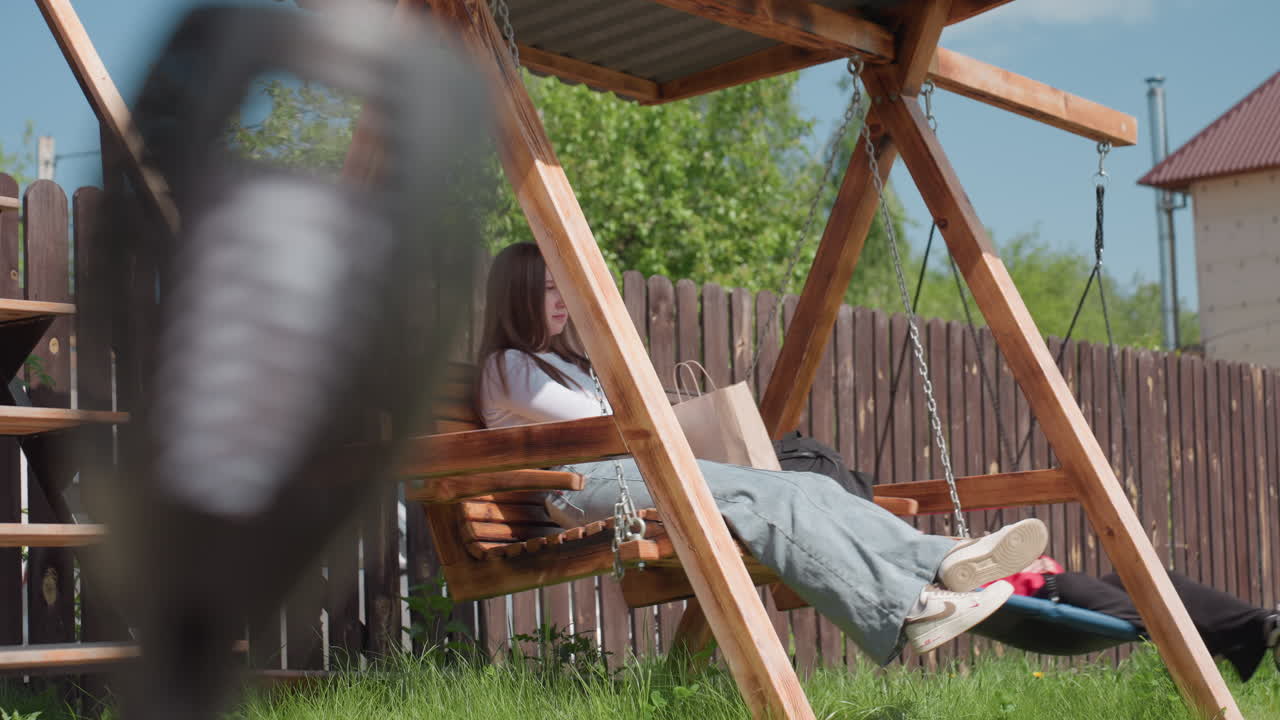 Woman sits calmly on wooden swing with shopping bags beside her while another person enjoys spinning on round swing under hot sun in cozy fenced backyard surrounded by lush greenery