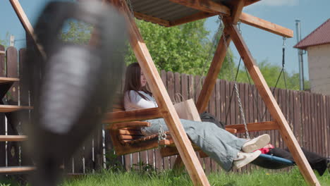 woman sits calmly on wooden swing with shopping bags beside her while another person enjoys spinning on round swing under hot sun in cozy fenced backyard surrounded by lush greenery