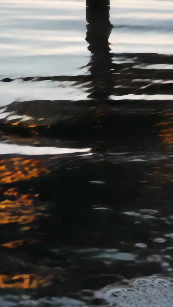 Vertical video: Wind stirring water, vertical post reflection shifting on dock at sunset with foam