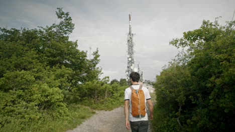 Camera-tracking-a-hiker-with-an-orange-backpack-walking-leisurely-towards-the-radio-tower-on-a-rocky-path-surrounded-by-high-green-gras-and-trees