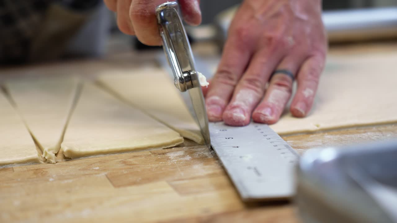 Dough rolls up on pastry wheel cutter as baker precisely measures and cuts dough into triangles for croissants, slow motion close up 4K
