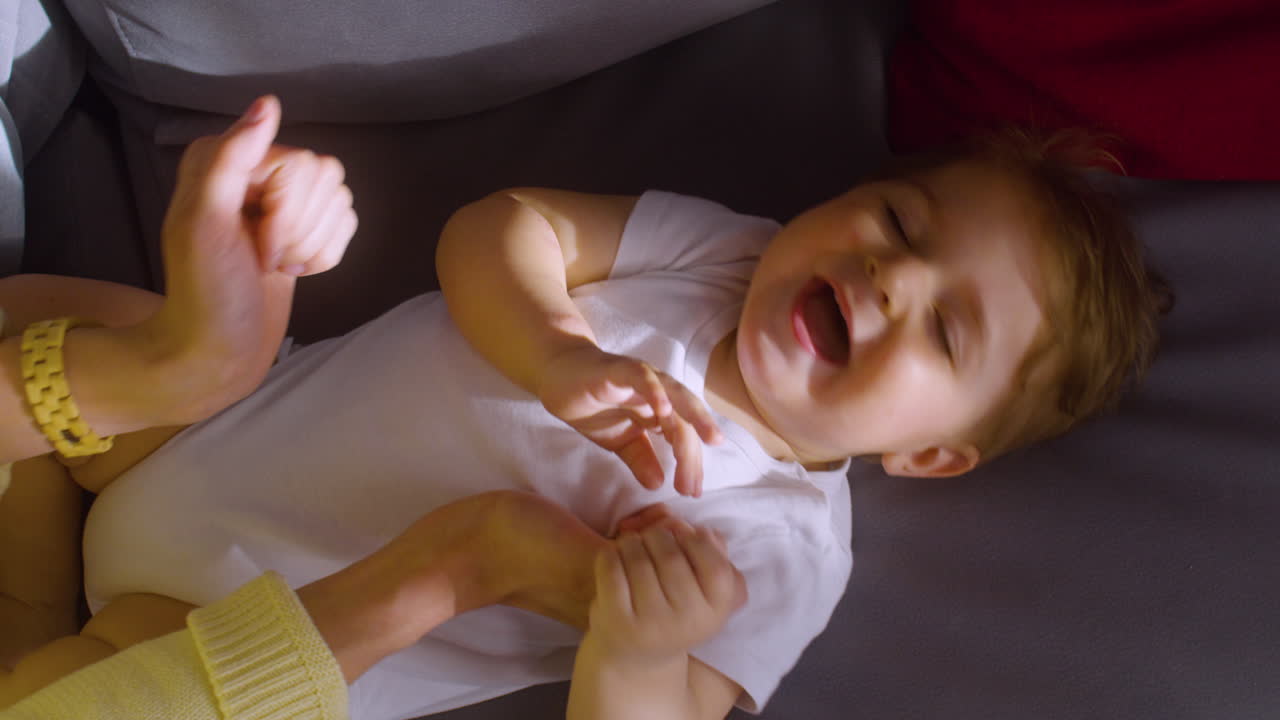 vista superior de un bebé sonriente acostado en el sofá en la sala de estar en casa mientras su madre le hace cosquillas y juega con él