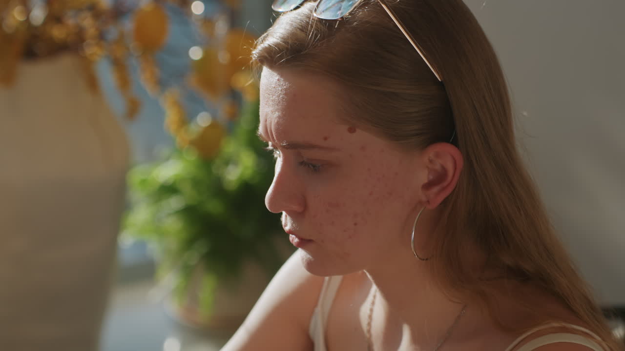 White woman pensive closeup at table, writer paused in thought, freckles and hoop earrings highlighted by warm sunlight, sunglasses perched on head, hand touching lips and temple, indoor plant