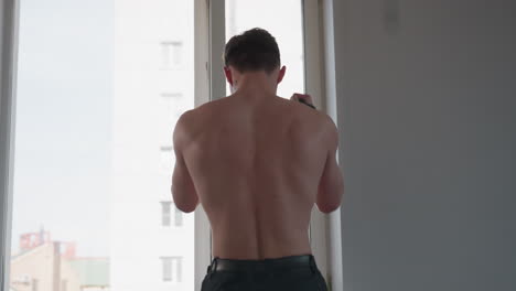 muscular shirtless young man pulling resistance rope while standing in front of large window, minimal interior with soft daylight, training session focused on back strength and upper body workout