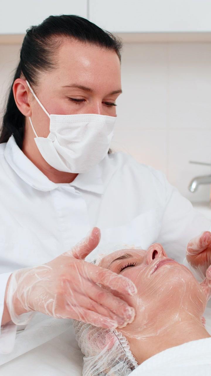 Beautician hands doing facial massage peeling serum on young adult woman relaxing in spa clinic