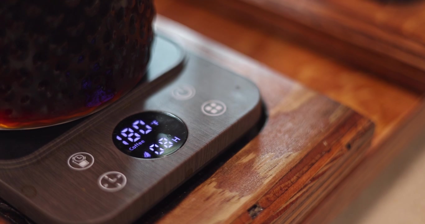 Barista adjusts digital temperature adjustment heater beneath a glass jar, coffee brewing on wooden table
