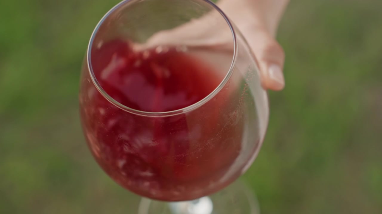 Una mano tiene un bicchiere pieno di liquido rosso, probabilmente vino o succo, contro uno sfondo esterno. L'attenzione rimane sulla texture e sul colore della bevanda, enfatizzando la sua ricca tonalità e chiarezza, sotto una luce soffusa.