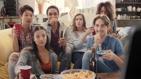 Diverse-group-of-student-friends-arms-raised-celebrating-goal-watching-sports-event-on-TV-together-eating-snacks--drinking-beer