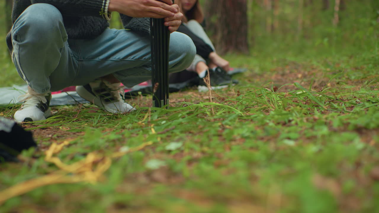 Partial view of person squatting and adjusting tent pole touching forest ground while another camper sits behind, slightly blurred, during calm camping setup in woodland environment