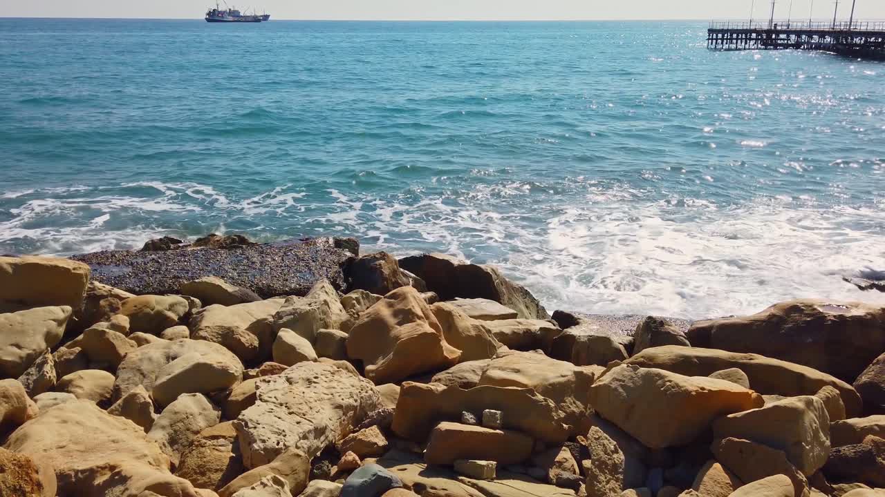 Ön planda büyük kayalıkların olduğu kıyıya vuran mavi deniz dalgaları. Güneş ışığı. Ağır çekim. Limasol, Kıbrıs