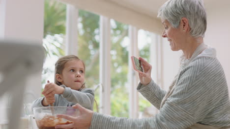 happy grandmother using smartphone taking photo of cute grandaughter in kitchen mixing ingredients for baking granny sharing weekend with grandchild on social media