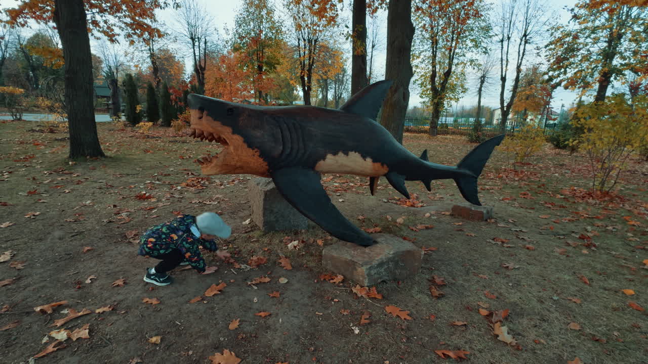 Little kid stands at the wooden sculpture of shark in the park. Baby boy picks something on the ground.
