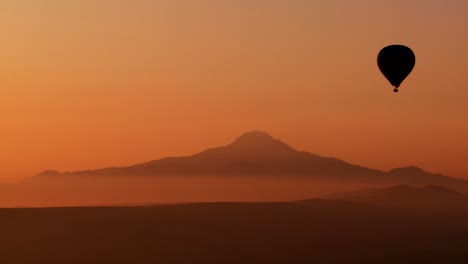 Hermosa-Toma-Aérea-De-Un-Globo-De-Aire-Caliente-Siluetas-Contra-Montañas-Distantes-En-Capadocia,-Turquía