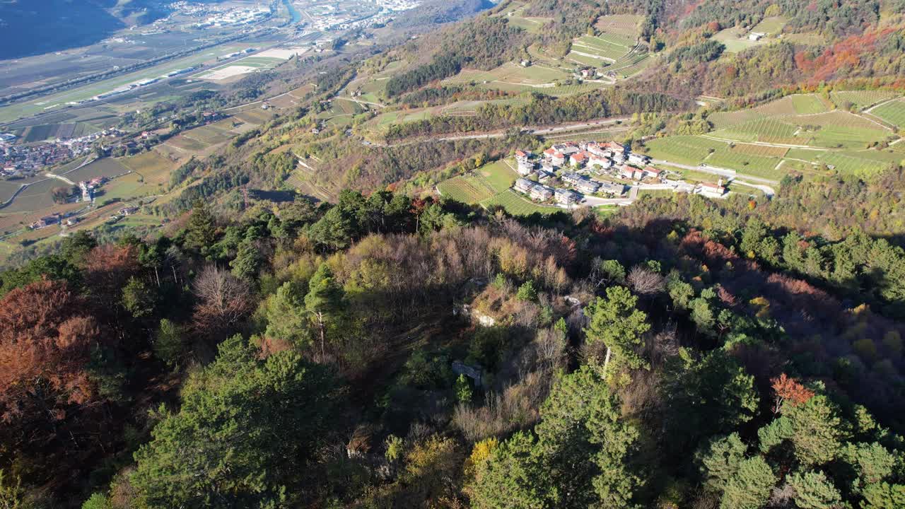 Cinematic aerial view of a small village on the mountains between vineyards, in Northern Italy in autumn. Trento City in the background