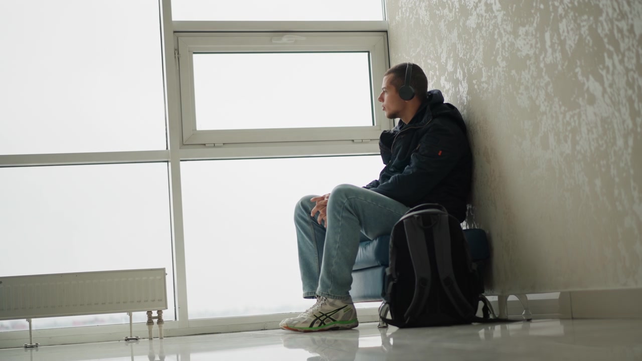 Hombre sentado tranquilamente en un banco cerca de grandes ventanales, usando auriculares y ropa casual, con equipaje a su lado. La escena es en interiores bajo iluminación suave, con paredes lisas y un fondo minimalista.
