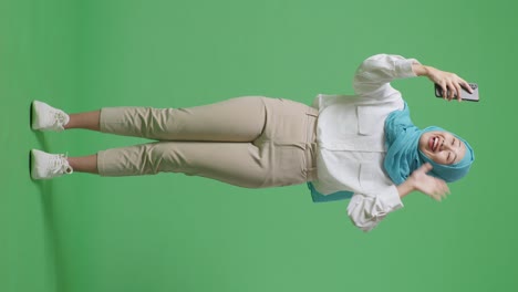 cuerpo lleno de una mujer musulmana asiática sonriente agitando la mano y haciendo una llamada de video en el teléfono inteligente mientras está de pie en el estudio de fondo de la pantalla verde