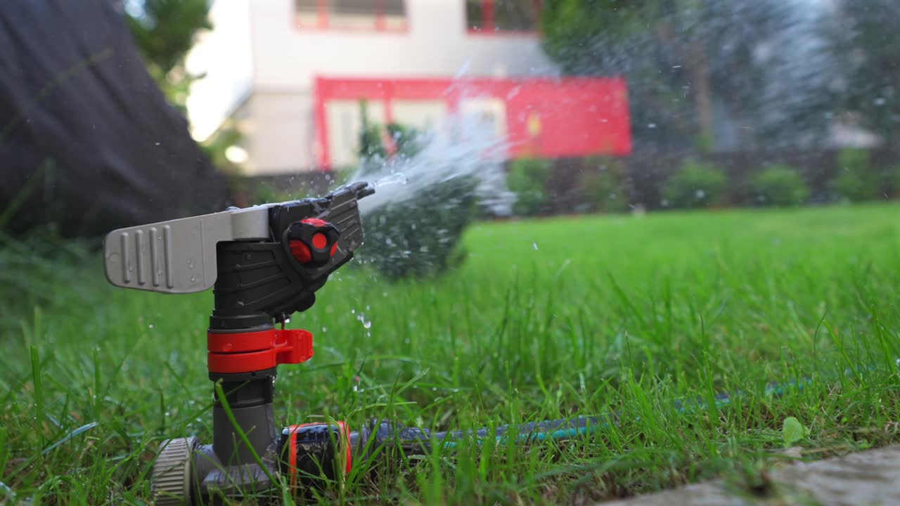 Orange gray impulse sprinkler in a lawn corner with green tube hose watering the lawn, a building and a construction container in the background, view from behind very close to the ground, closeup