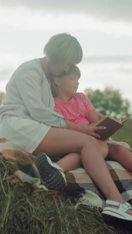 una madre abrazando a su hija mientras se sienta en el follaje del heno al anochecer, leyendo juntos, con un sombrero cerca, rodeados por un campo abierto
