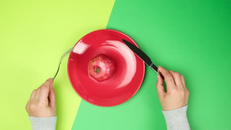 red ceramic plate with red ripe pomegranate and two female hands holding a metal knife and fork