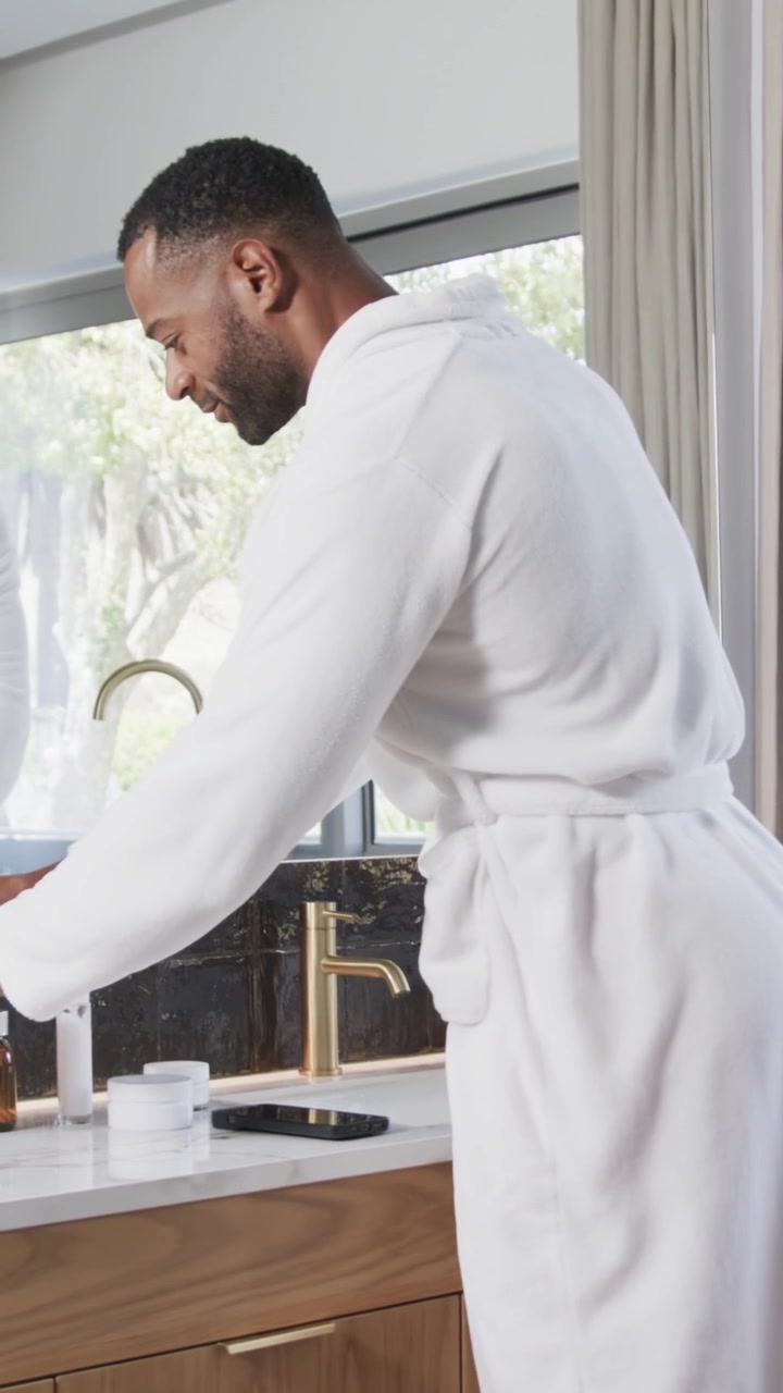 Vertical video: Answering call, man in robe talking on phone at sink, applying cream for grooming