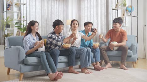 아시아 십대들이 집에서 스마트폰으로 축구 경기를 보는 동안 환호하고 먹고 마시고 있습니다.