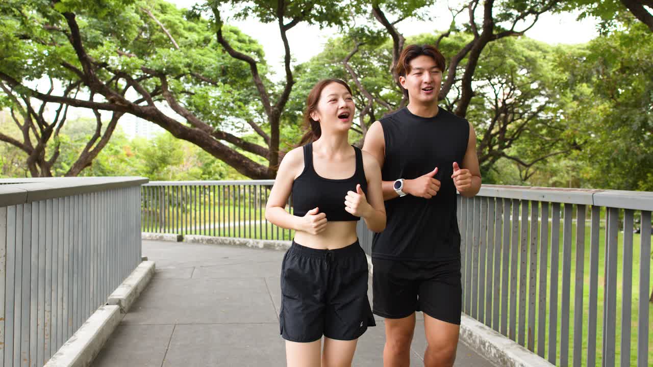 Coppia asiatica sorridente che fa jogging su un ponte in un parco verde della città, ripresa media a mano libera, luce naturale
