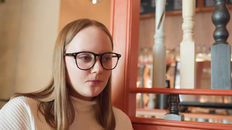 young woman in glasses leans against wooden railing in cozy indoor cafe, gazing calmly with satisfied expression after meal, surrounded by soft light, colorful decor, and blurred background