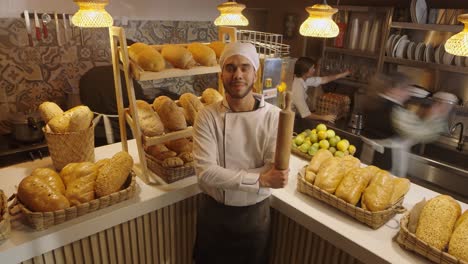 a smiling baker in a white uniform stands proudly in front of his pastries, rolling pin in hand.