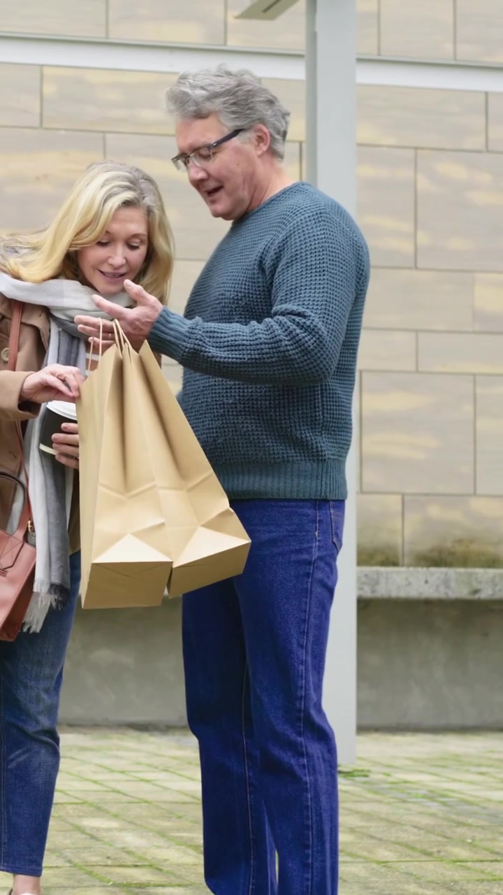 Video verticale: Un uomo in abiti color verde acqua in piazza mostra il telefono e le borse a una donna che si avvicina con una tazza.