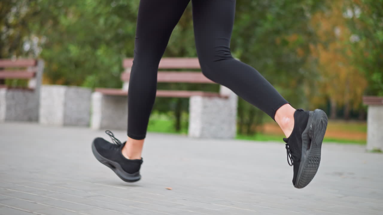 Close-up view of woman's legs in black leggings and sneakers running in park path, showing motion, fitness routine, and outdoor jogging with park bench and nature in background
