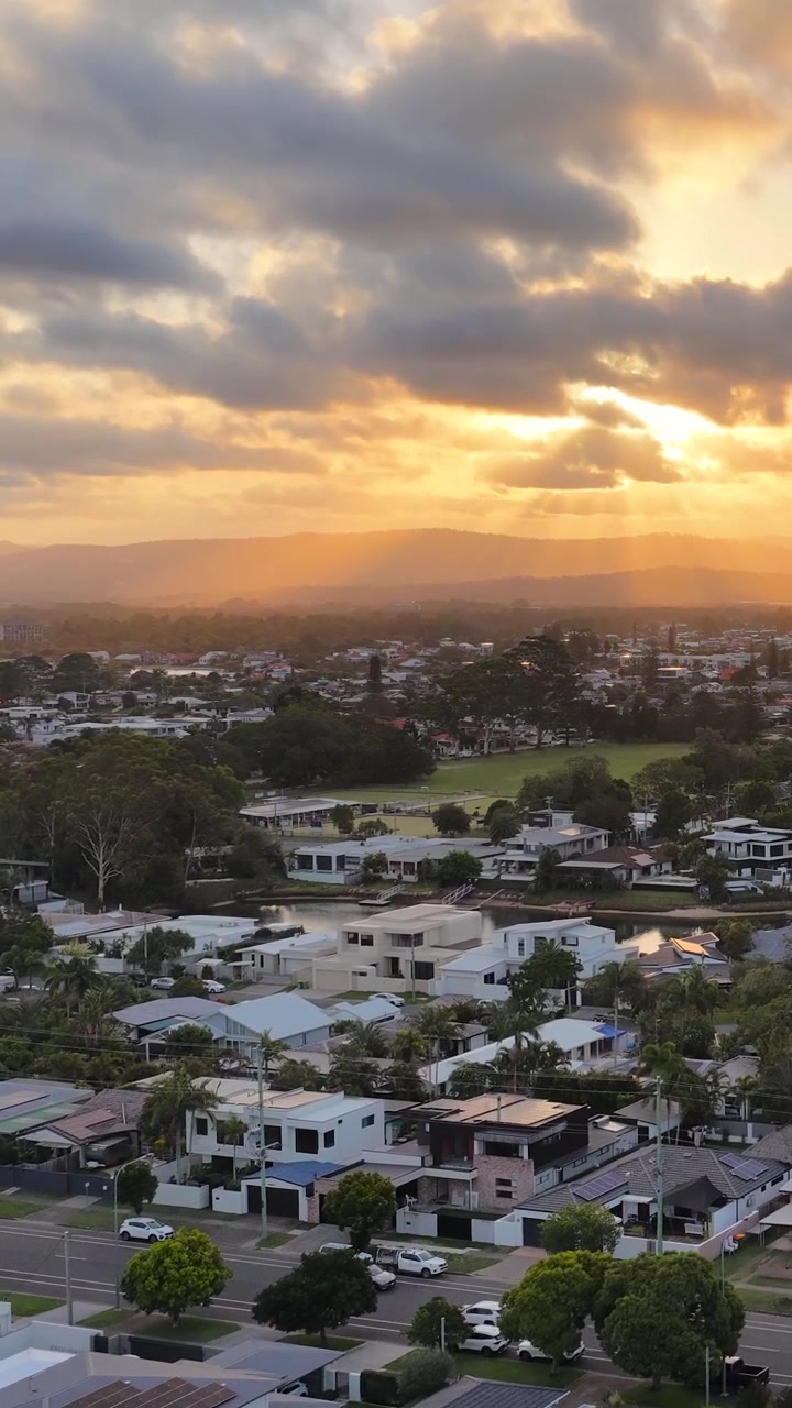 Un dron se desliza sobre suburbios frente al mar al atardecer, revelando casas de lujo, canales y un cielo espectacular.