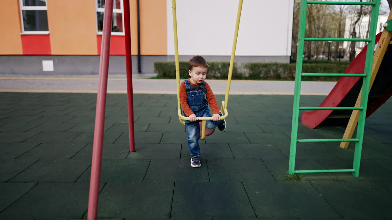 Lovely boy sits on a swing swaying slowly and pushing himself. Two-year-old toddler spending time at the playground.
