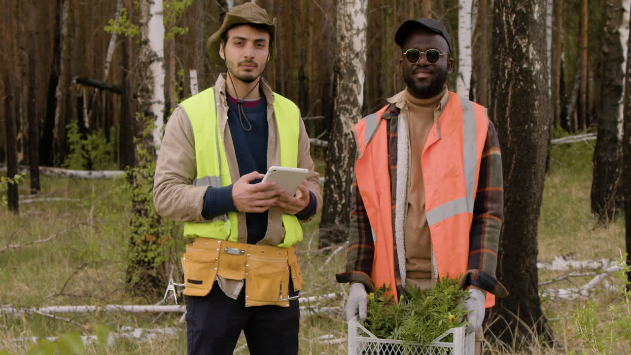 activistas caucásicos y afroamericanos sostienen una tableta y pequeños árboles mientras miran la cámara en el bosque
