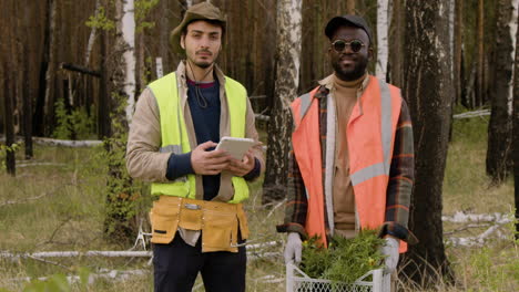 caucasian and african american men activists holding a tablet and small trees while looking at camera in the forest