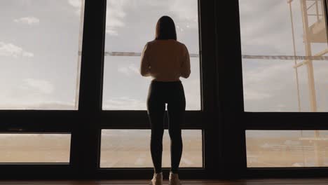 Low-angle-approach-toward-female-stand-near-wide-studio-apartment-window