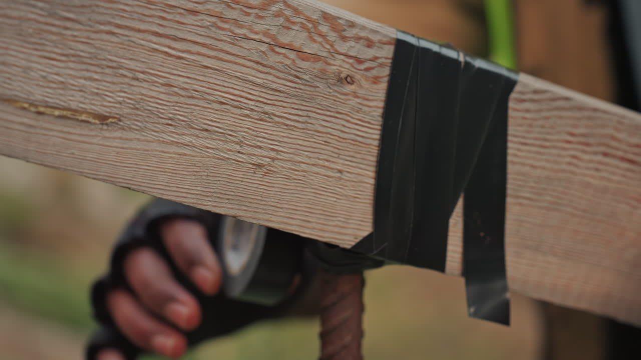 Primo piano del processo di riparazione di un'asse di legno con nastro adesivo, mani guantate che avvolgono nastro telato nero attorno all'asse e al tondino arrugginito, movimenti precisi e costanti, cantiere esterno, venatura del legno, aspetto pratico.