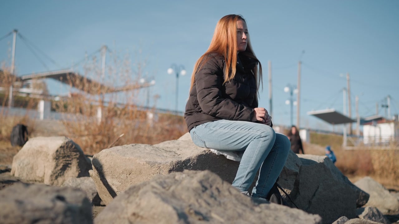 Woman sits quietly on large rocks in outdoor setting wearing black jacket and light blue jeans. background features distant structures under clear skies with soft daylight illuminating scene