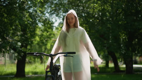 young woman in translucent raincoat stands near bicycle with arms outstretched, joyfully swinging her hands as if wanting to fly, background filled with lush greenery