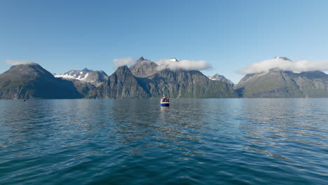 Drone-flying-at-low-altitude-toward-fishing-boat-navigating-in-Lyngen-fjord-with-rocky-mountains-of-Scandinavian-Alps-in-background,-Norway