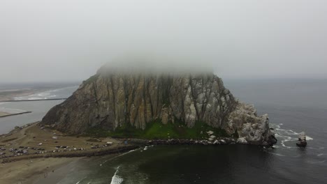 counterclockwise drone shot of morro rock covered by fog on a cloudy day in morro bay, california. the sea stack towers over the ocean waves and the beach below as the cliffs contrast greenery.