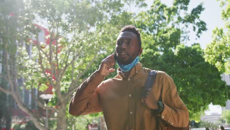 hombre de negocios afroamericano con máscara facial hablando por teléfono inteligente en la calle