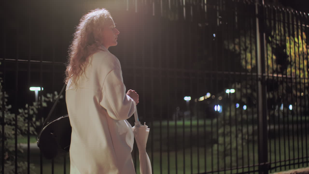 Young lady walking along iron fence under streetlights, looking right with soft light shining on curly hair, blurred bokeh lights glowing in background