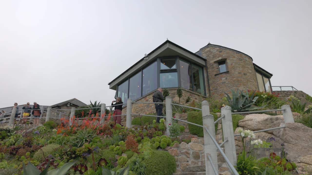 People exploring a terraced coastal garden with stone steps and buildings under an overcast sky