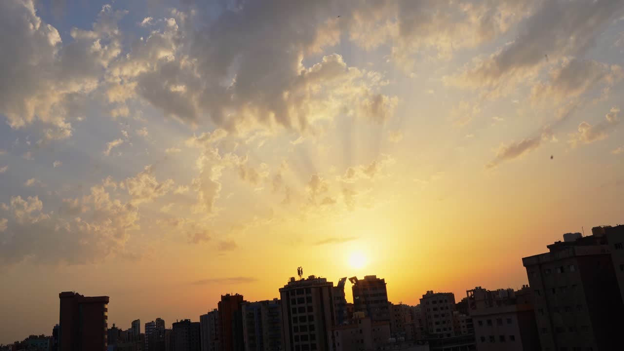 Plano geral de um pôr do sol dourado e quente sobre o horizonte denso de uma cidade. Raios de sol atravessam nuvens dispersas sobre arranha-céus durante a mágica hora dourada em uma paisagem urbana.