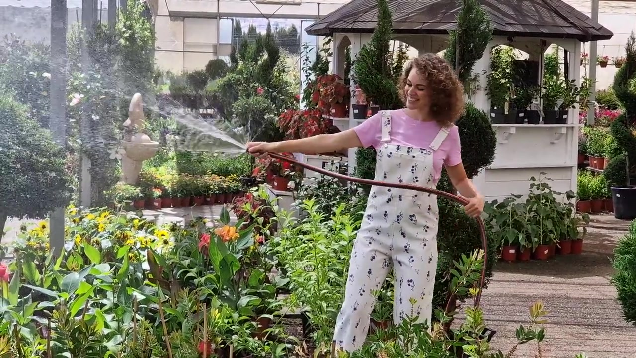 Giovane donna felice in piedi che innaffia le piante con la canna dell'acqua in un giardino verde.