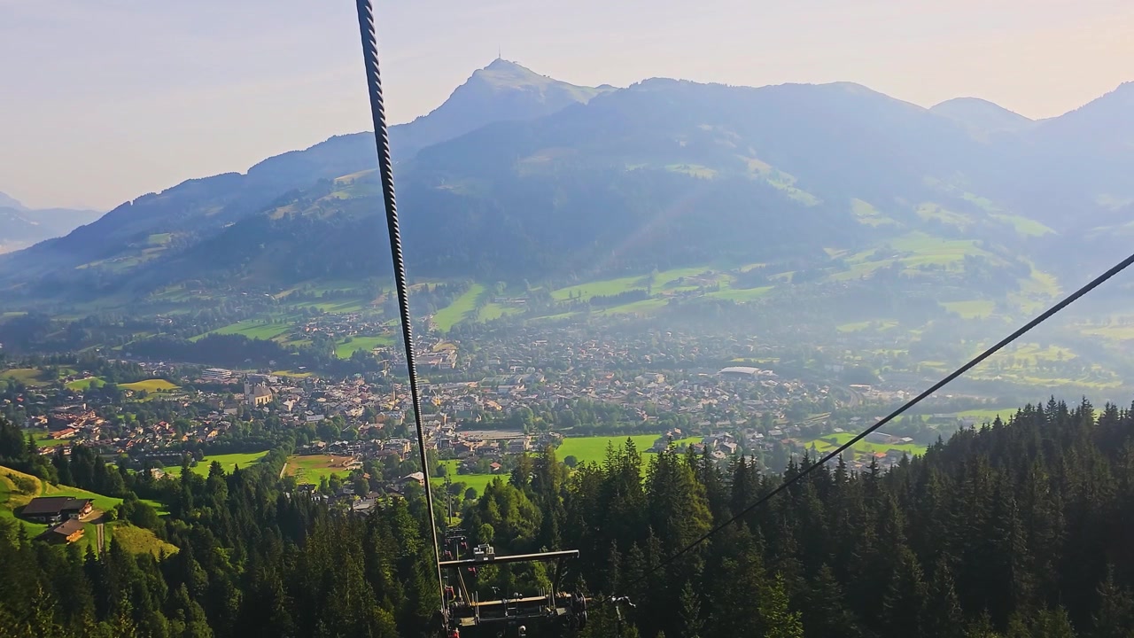 Funivia che sale sopra Kitzbühel, Austria, con cabine rosse, case chalet, pascoli verdi, fitta foresta di pini e creste montuose con tempo estivo luminoso in una vista panoramica del Tirolo
