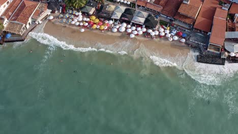 rising aerial drone birds eye shot of the beautiful tropical famous pipa beach during high tide with tourists playing in the water and enjoying the shade under colorful umbrellas on a summer evening