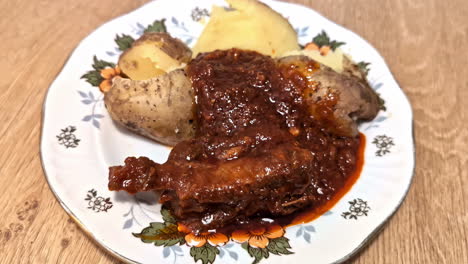 boiled jacket potatoes with chicken wings in tomato sauce. homemade dinner on a white plate with a decorative pattern. chicken in sauce garnished with potatoes.