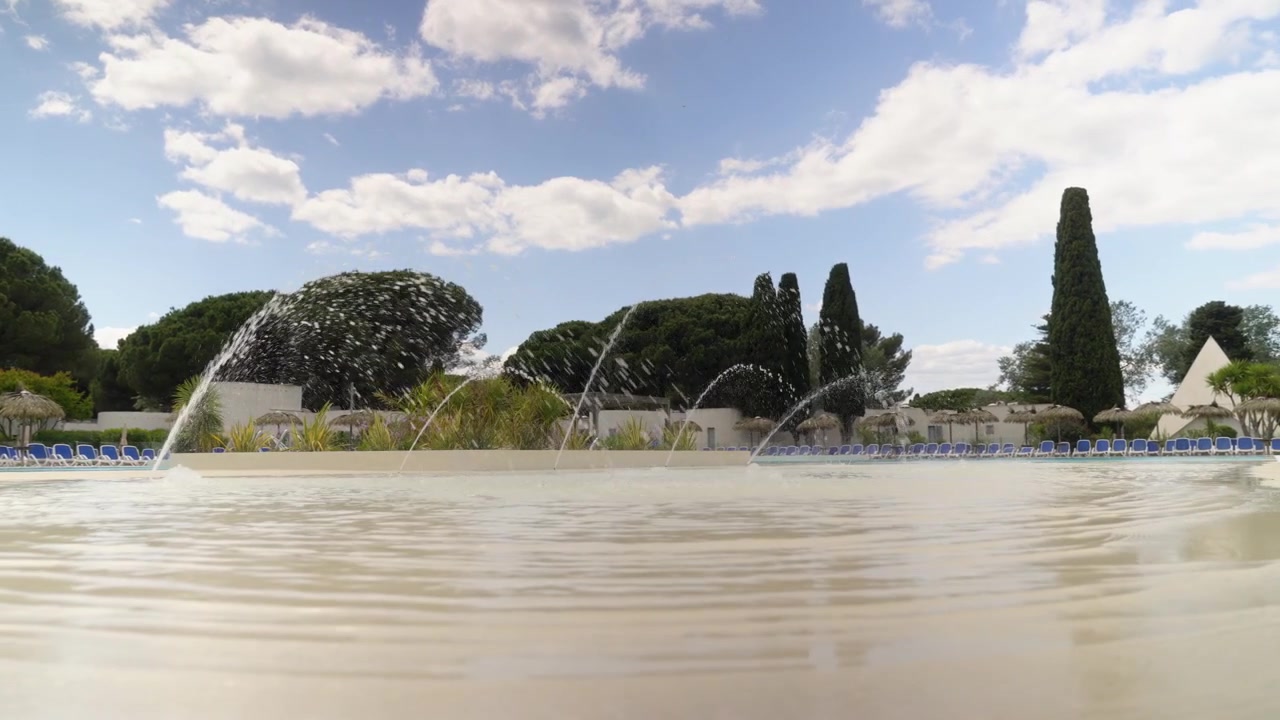 Una vista a basso livello dell'acqua mostra getti di fontane ad arco che spruzzano in una piscina all'aperto presso un resort, con alberi e un cielo azzurro parzialmente nuvoloso sullo sfondo