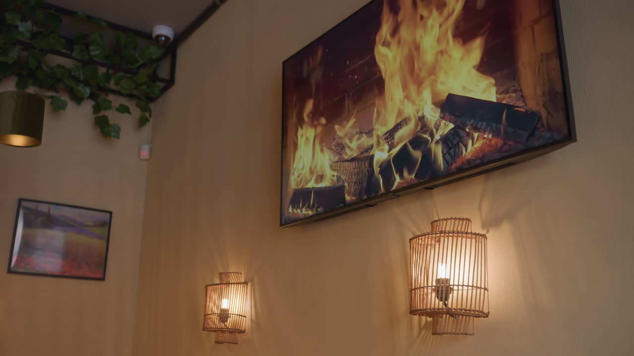 Beautiful view of glowing light bulbs enclosed in wooden decorative frames on wall beside television displaying animated fireplace flame, creating warm cozy ambiance with indoor greenery